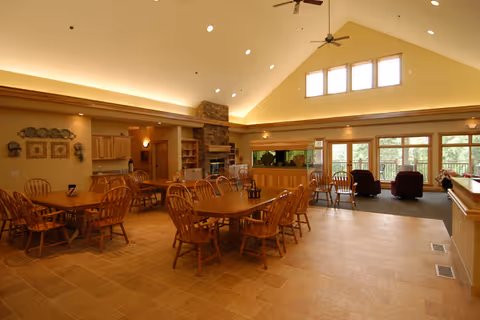 A spacious, well-lit common area in an assisted living facility featuring multiple wooden dining tables with chairs, a stone fireplace, large windows letting in natural light, and a seating area with armchairs near the windows. The ceiling is vaulted with ceiling fans and recessed lighting.