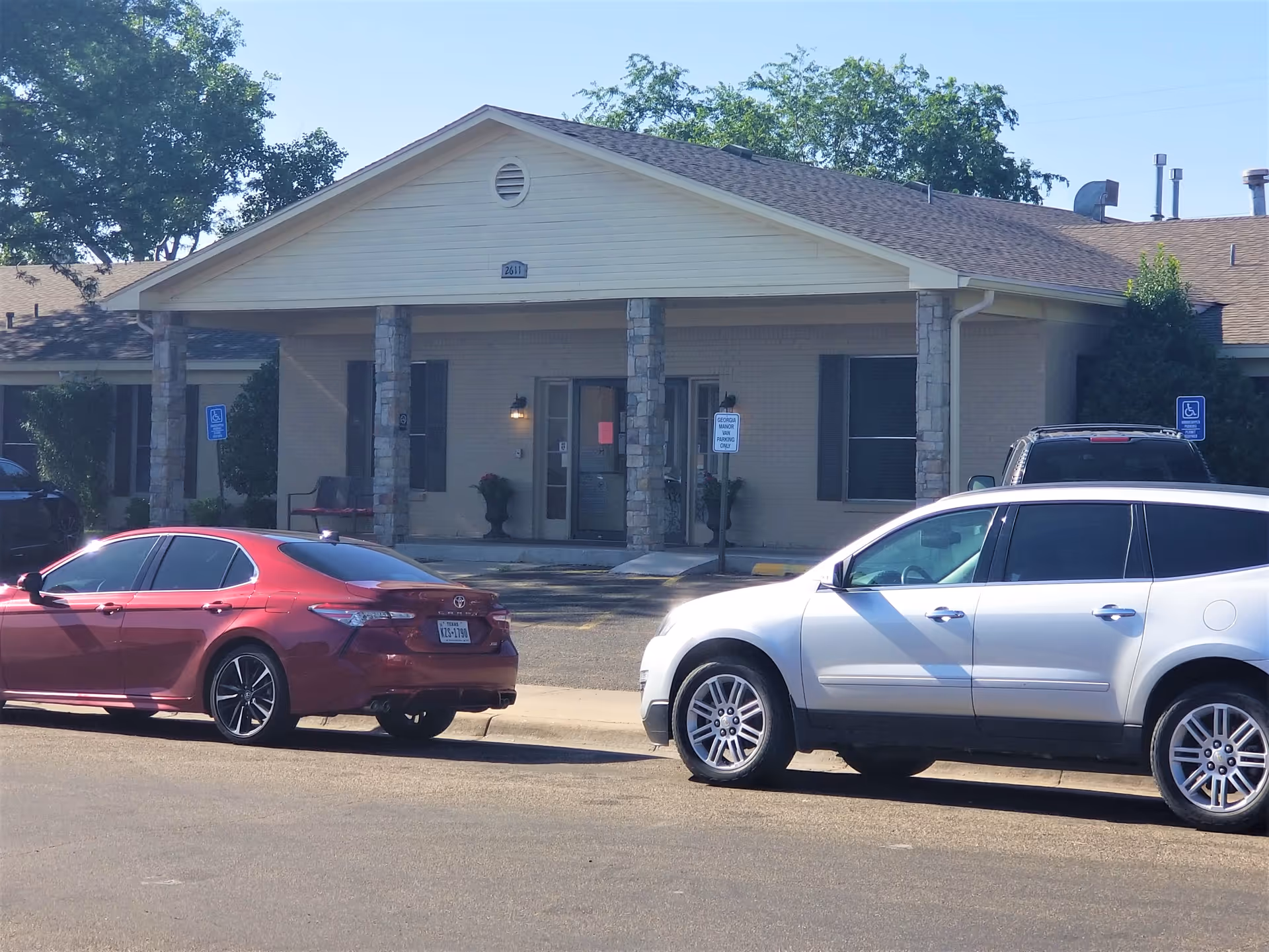 Front exterior view of Georgia Manor Nursing Home building with a covered entrance supported by stone columns. Two cars are parked in front, and there are signs indicating designated parking spaces. Trees and clear sky are visible in the background.