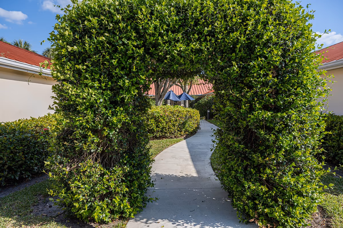 A concrete pathway passes through a trimmed leafy archway into a courtyard between single-story buildings with red roofs and patio umbrellas.