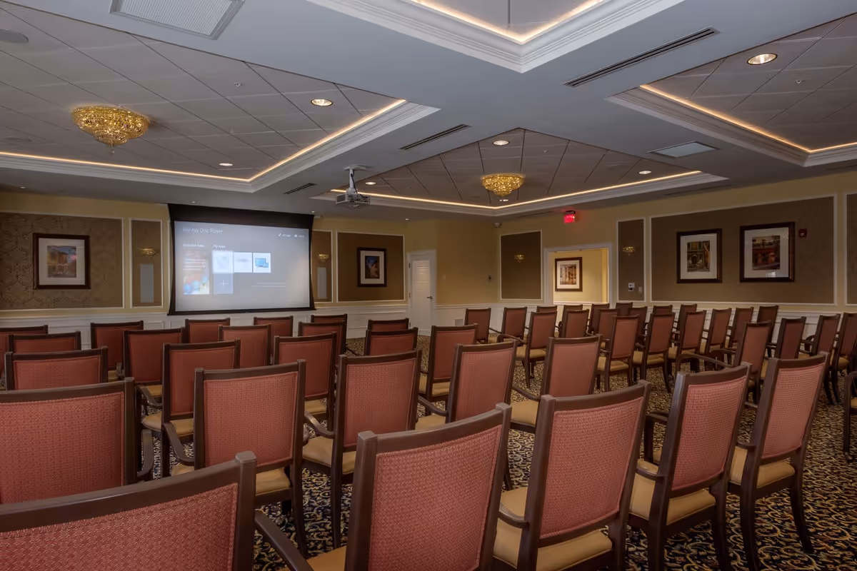 Empty presentation/meeting room with rows of chairs facing a projector screen under decorative ceiling lights.