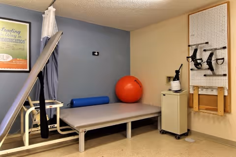 A small rehabilitation room featuring a treatment table, a red exercise ball, a blue foam roller, a tilt table, and therapy equipment mounted on a pegboard.