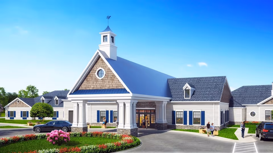 Front entrance of a low‑rise senior living building with a tall pitched roof, columns, landscaping and parked cars.
