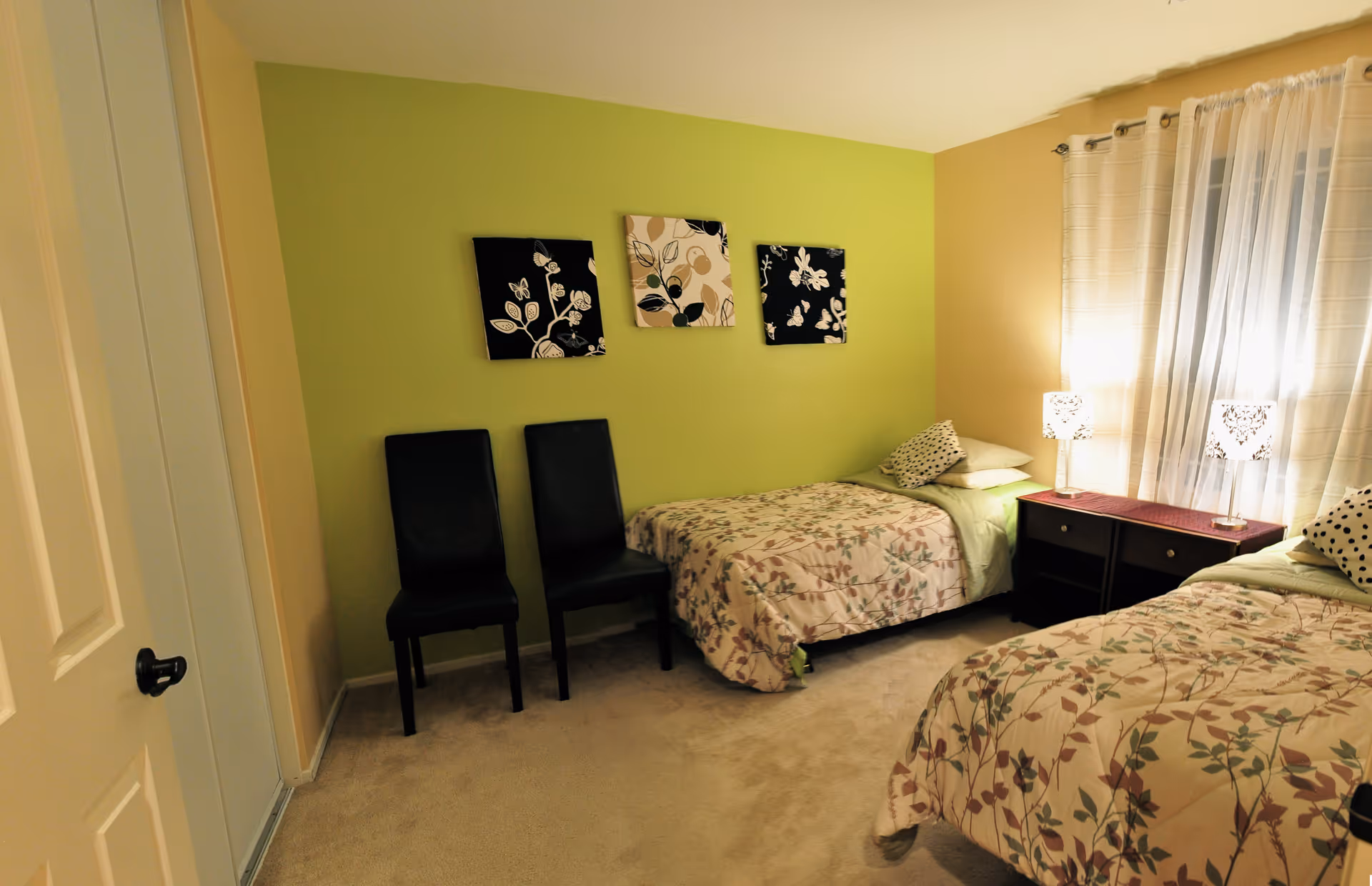A cozy bedroom with two single beds covered in floral patterned bedspreads. Between the beds is a dark wooden nightstand with two decorative lamps. The walls are painted green and beige, with three floral-themed artworks hanging on the green wall. Two black chairs are placed against the green wall near a closed white door. A window with sheer white curtains allows soft light into the room.