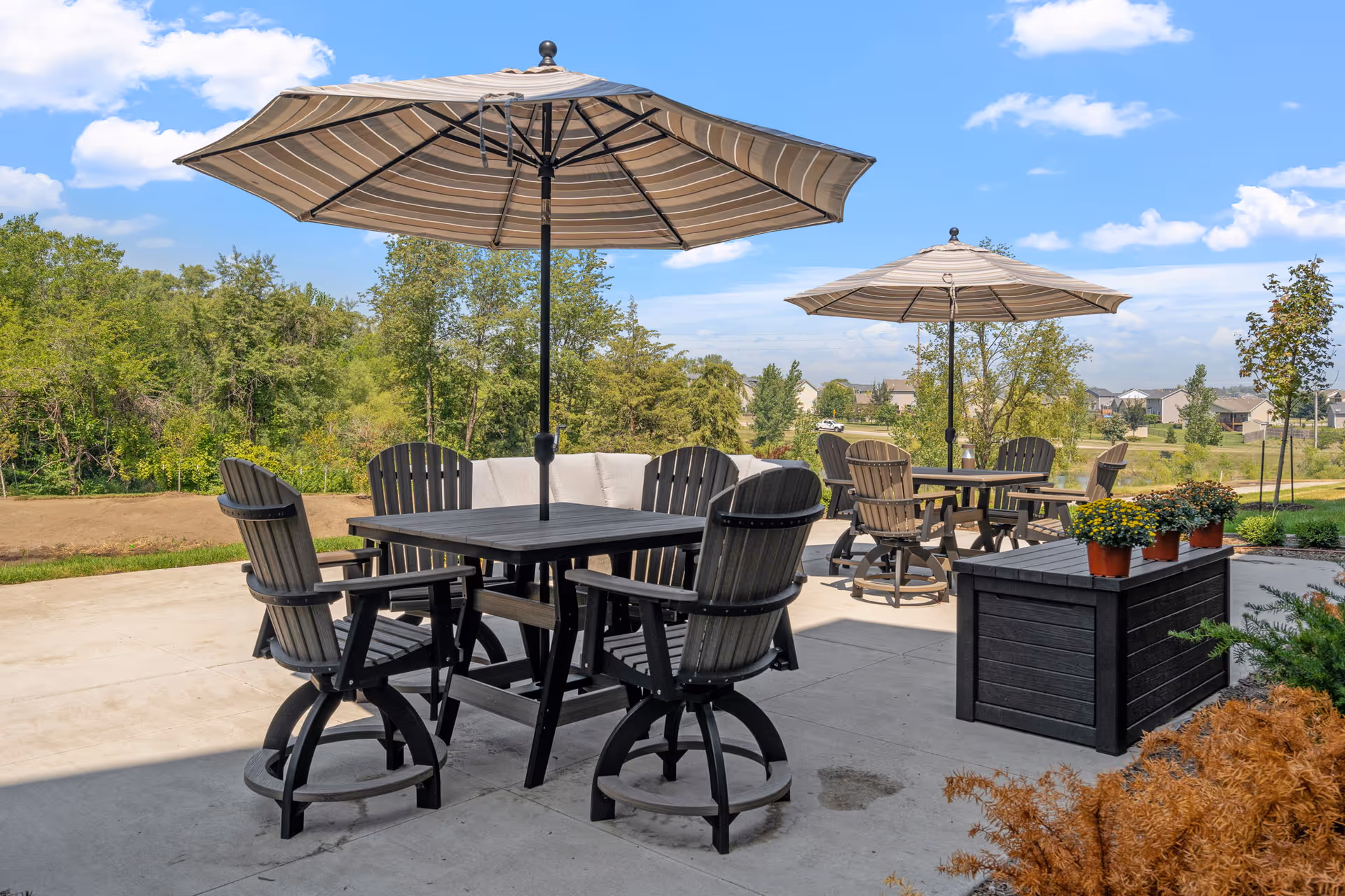 Patio dining area with tables, striped umbrellas, wooden chairs and potted plants overlooking trees and nearby homes.