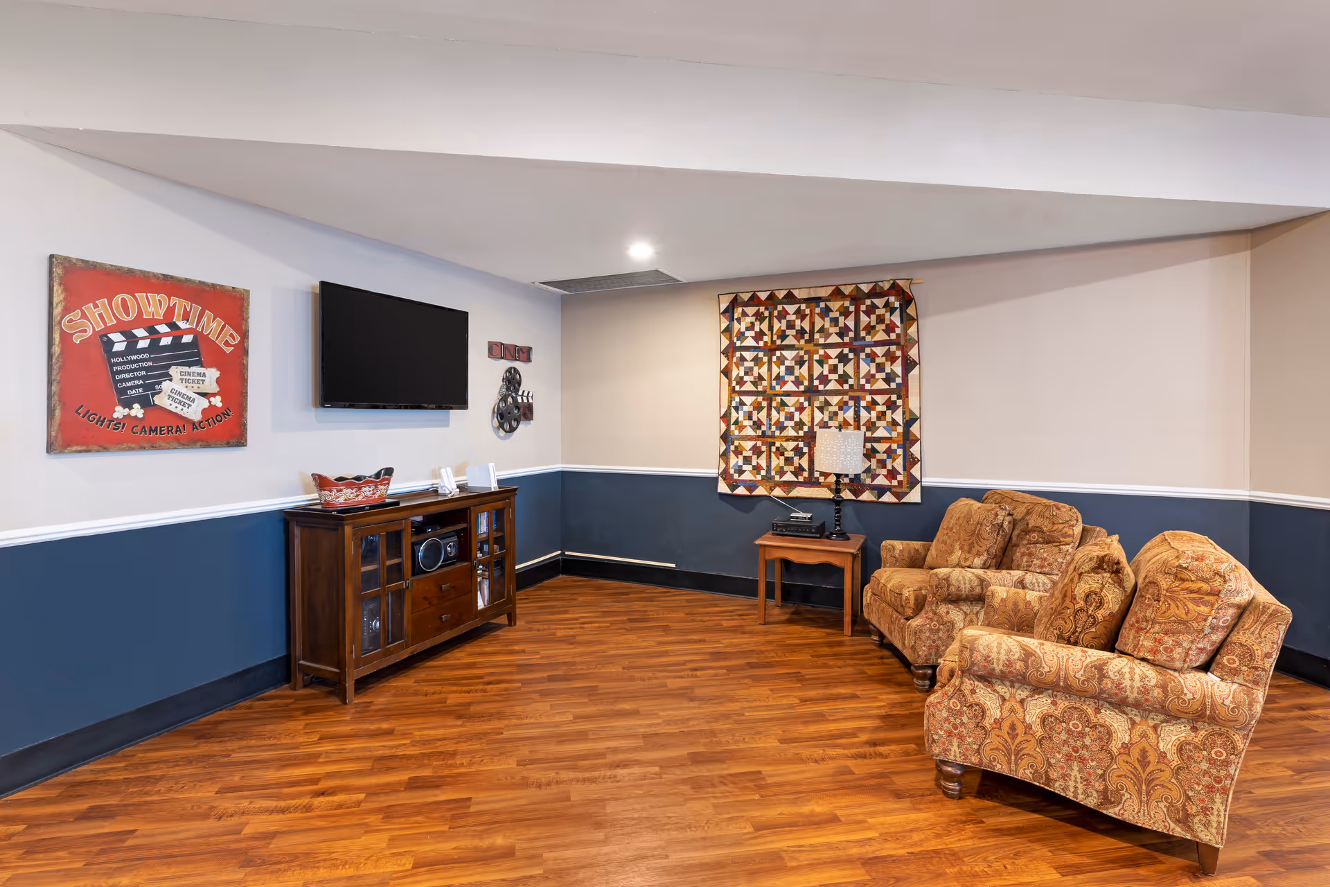 A cozy living room area with two patterned armchairs and a small wooden side table with a lamp. A colorful quilt hangs on the wall above the side table. On the adjacent wall, there is a flat-screen TV mounted above a wooden cabinet with glass doors and a decorative basket on top. A red wall art piece with the word 'SHOWTIME' and a movie clapperboard design is also visible. The room has wood flooring and walls painted in two tones with a white chair rail.