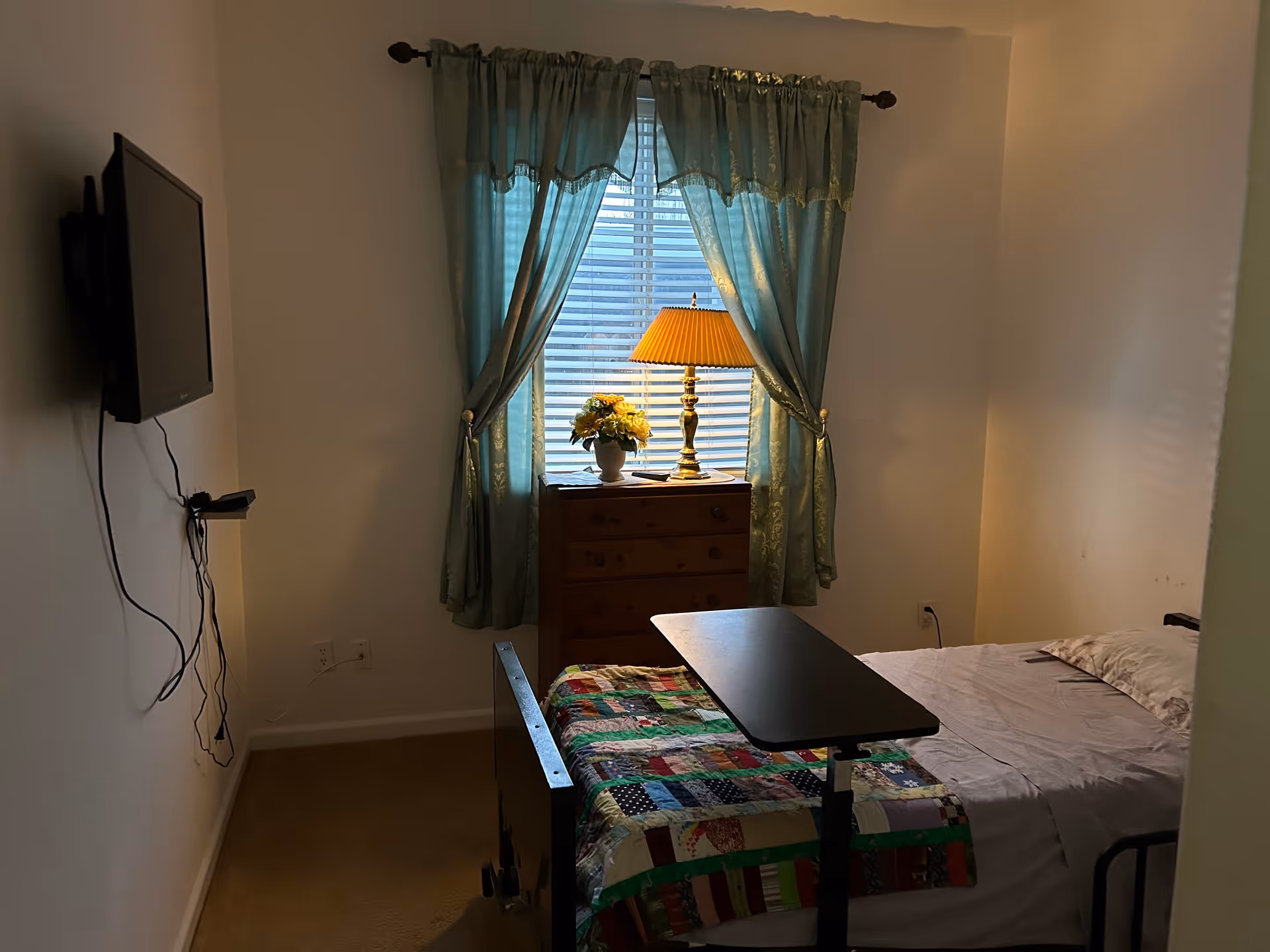 A small bedroom with a single bed covered with a colorful patchwork quilt and white sheets. There is a black overbed table positioned over the bed. A wooden dresser with a yellow lamp and a vase of flowers sits in front of a window with green curtains. A flat-screen TV is mounted on the wall to the left.
