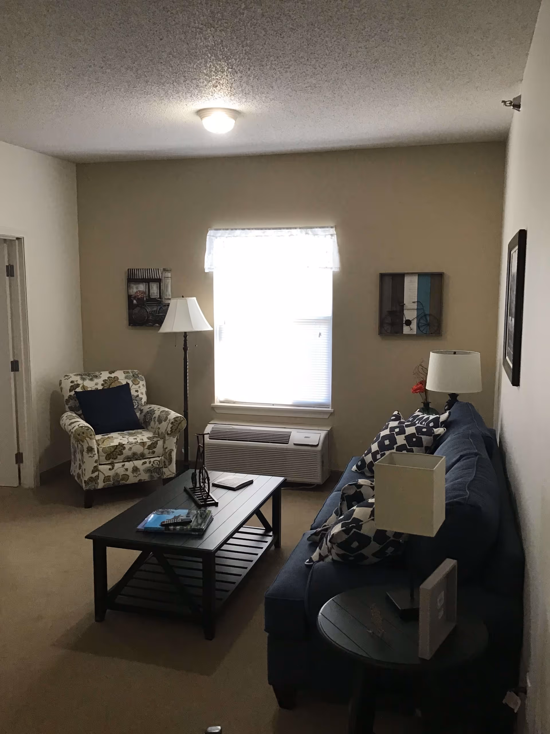 A cozy living room with a blue sofa adorned with patterned cushions, a floral armchair with a dark blue pillow, a black coffee table with books and decorative items, a floor lamp, and a window with a white curtain above an air conditioning unit. The walls are decorated with framed artwork.