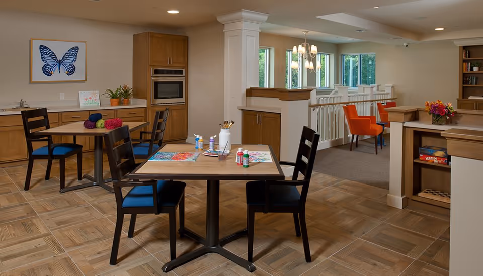 A well-lit activity room with wooden tables and chairs featuring blue cushions. One table has art supplies including paint bottles, brushes, and paintings, while another table has colorful yarn. The room has wooden cabinetry, a built-in oven, a large butterfly artwork on the wall, and a cozy seating area with orange chairs near windows overlooking greenery.