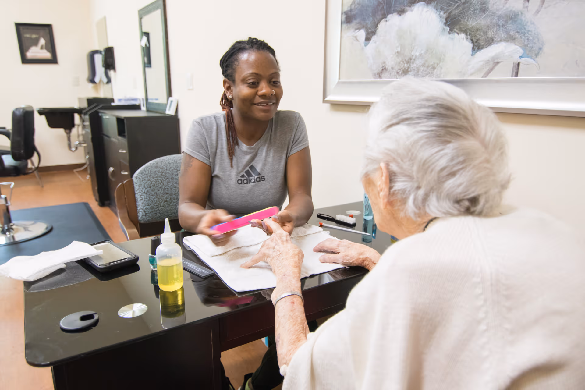 A young woman providing a manicure to an elderly woman seated across a black table in a well-lit room with salon chairs and a large framed artwork on the wall.