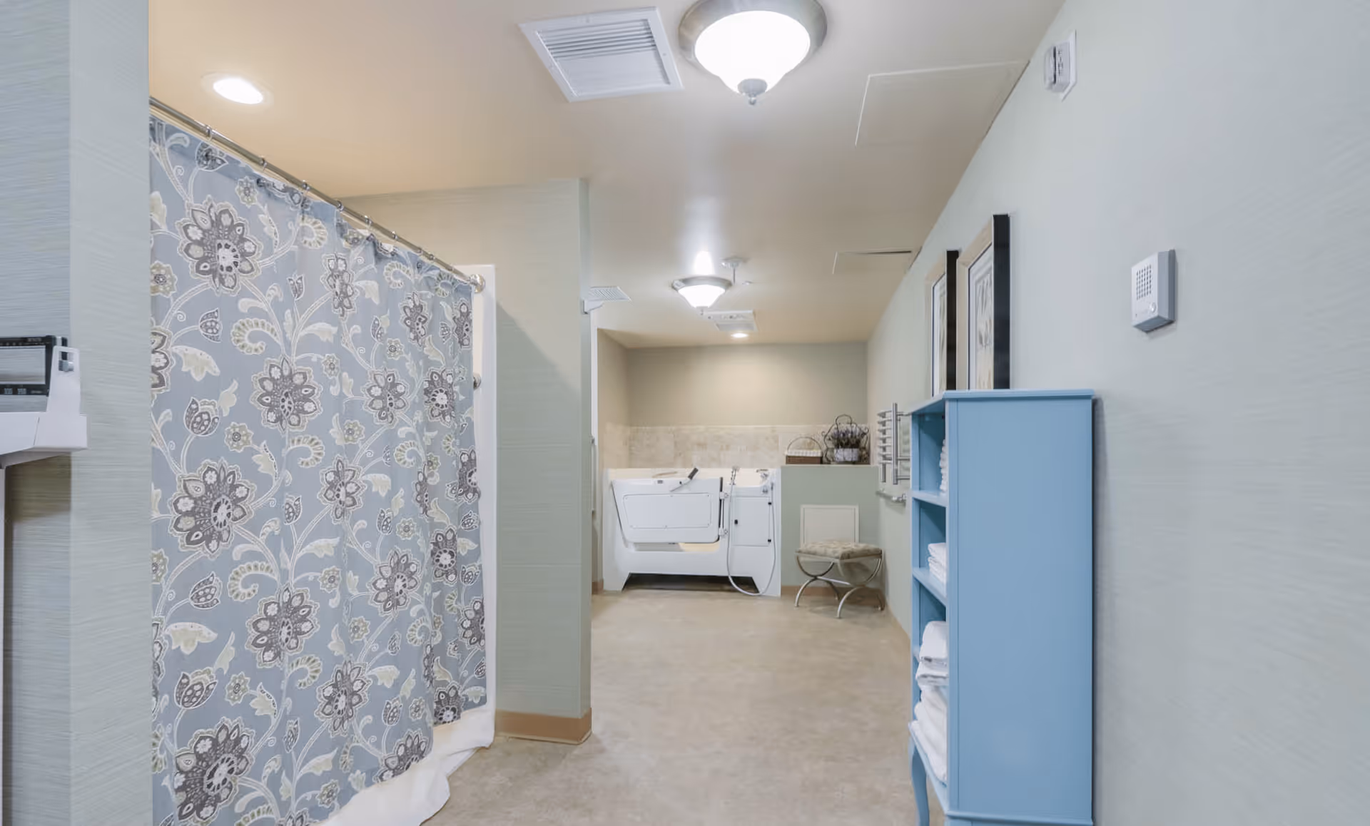 A spacious assisted living bathroom area featuring a walk-in bathtub with a floral shower curtain on the left, a blue shelving unit stocked with white towels on the right, and a small cushioned stool near the back wall. The room is well-lit with ceiling lights and has light-colored walls and flooring.