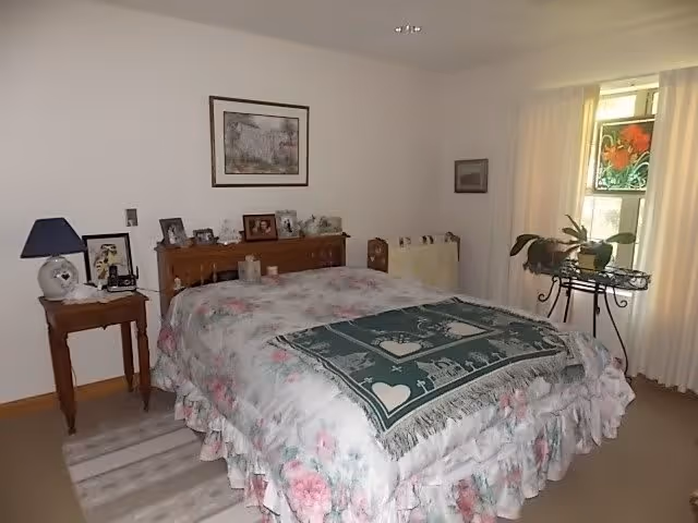 A bedroom with a bed covered in floral bedding and a green decorative throw. There is a wooden headboard with framed photos and small items on it. A small wooden side table with a lamp and a clock is next to the bed. A window with light curtains and a stained glass decoration is on the right side, with a plant on a metal stand nearby. The walls are light-colored and there is a framed picture hanging above the bed.