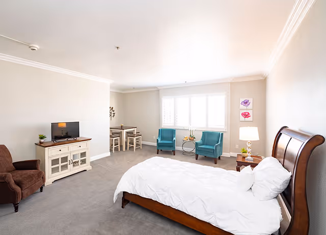 A bright and spacious bedroom in a retirement center featuring a wooden bed with white bedding, a brown armchair, a small TV on a white cabinet, two teal armchairs near a window with white shutters, a small round table with a plant, a wooden nightstand with a lamp and plant, and a small dining table with two chairs in the corner.