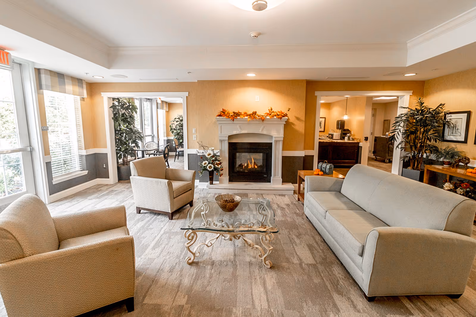 Bright furnished common living room with sofa, armchairs, glass coffee table, and a lit fireplace.