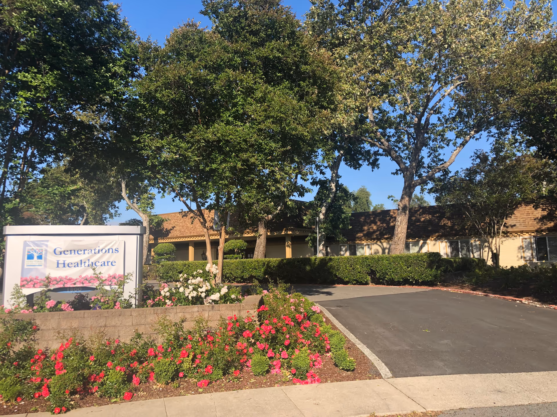 Exterior view of a healthcare facility with a sign reading 'Generations Healthcare' surrounded by blooming flowers and trees, with a driveway leading to the building under a clear blue sky.