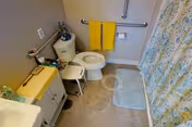 A small bathroom with a toilet, a white cabinet with a yellow towel on top, a white sink, a shower curtain with a floral pattern, a blue bath mat, and two yellow towels hanging on a grab bar above the toilet.