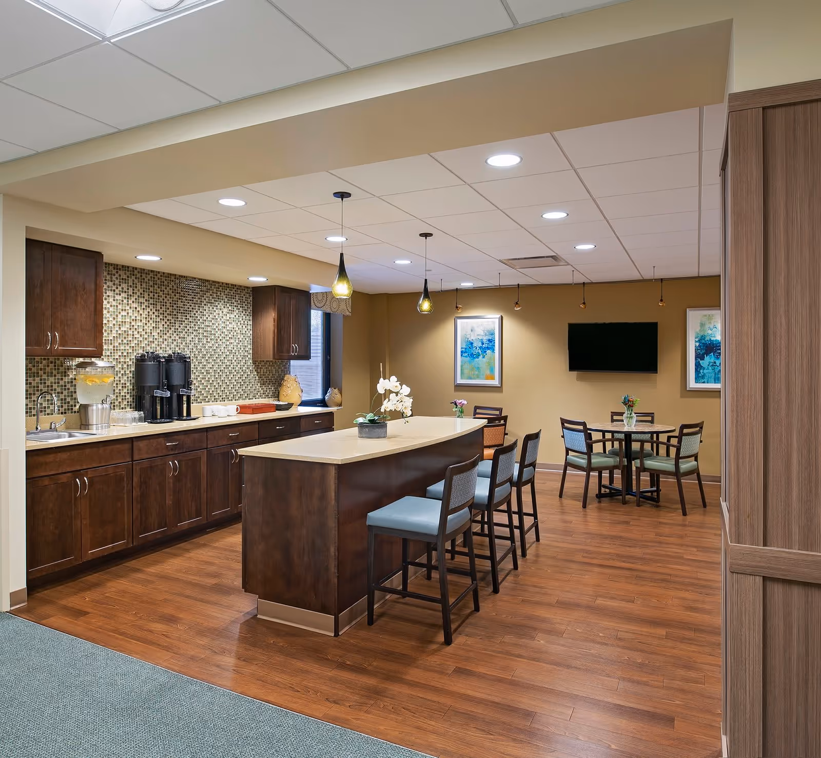 A modern common area with a kitchen and dining space. The kitchen features dark wood cabinets, a mosaic tile backsplash, a countertop with a water dispenser and coffee machines, and an island with four chairs. The dining area has a round table with four chairs, a wall-mounted TV, and two colorful framed artworks on the wall. The floor is wood, and the ceiling has recessed lighting and pendant lights above the island.