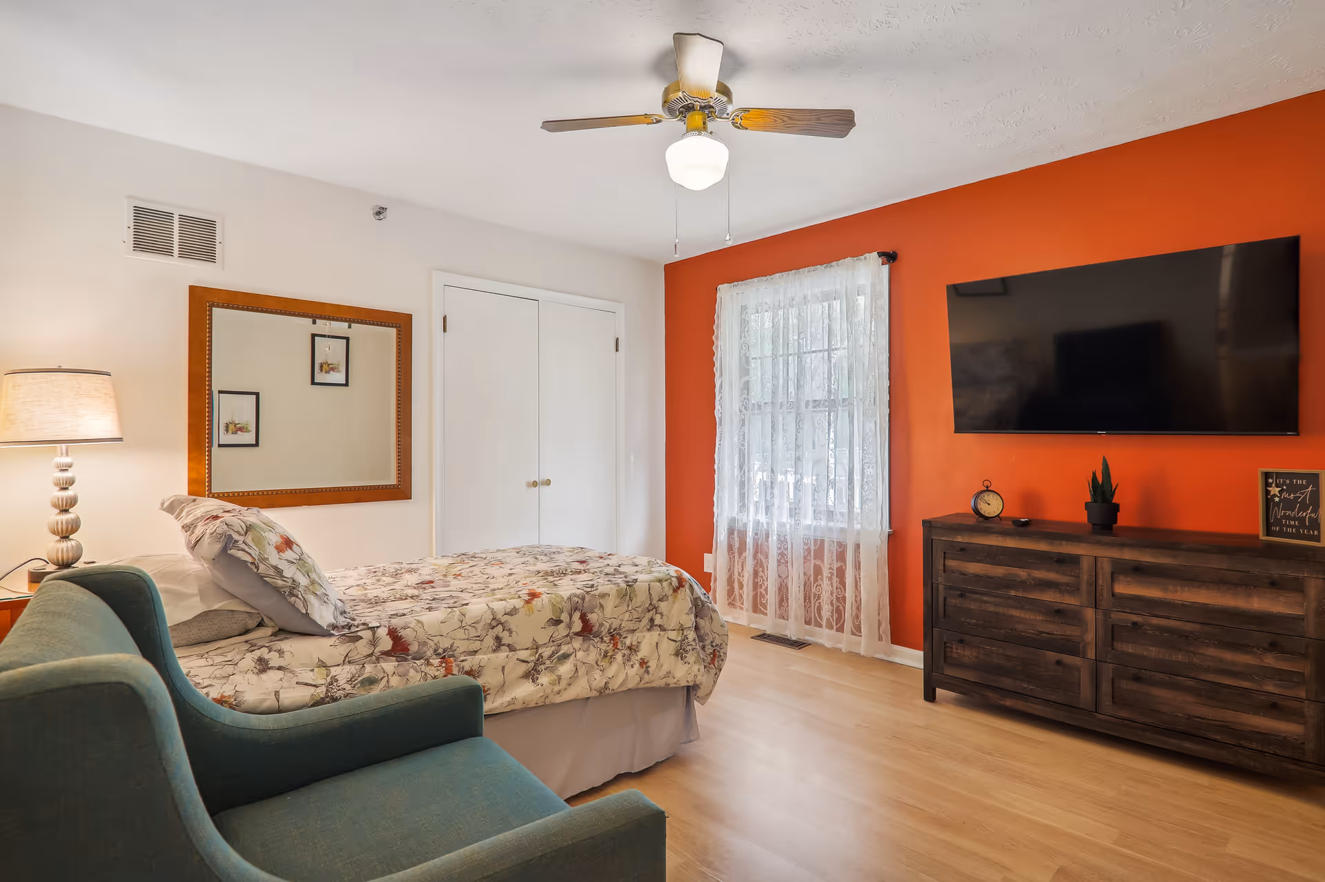 A cozy bedroom with a floral bedspread on a bed, a green armchair, a wooden dresser with a potted plant and a clock, a large flat-screen TV mounted on an orange accent wall, a window with sheer white curtains, a ceiling fan with a light, and a lamp on a bedside table next to a large mirror on the wall.