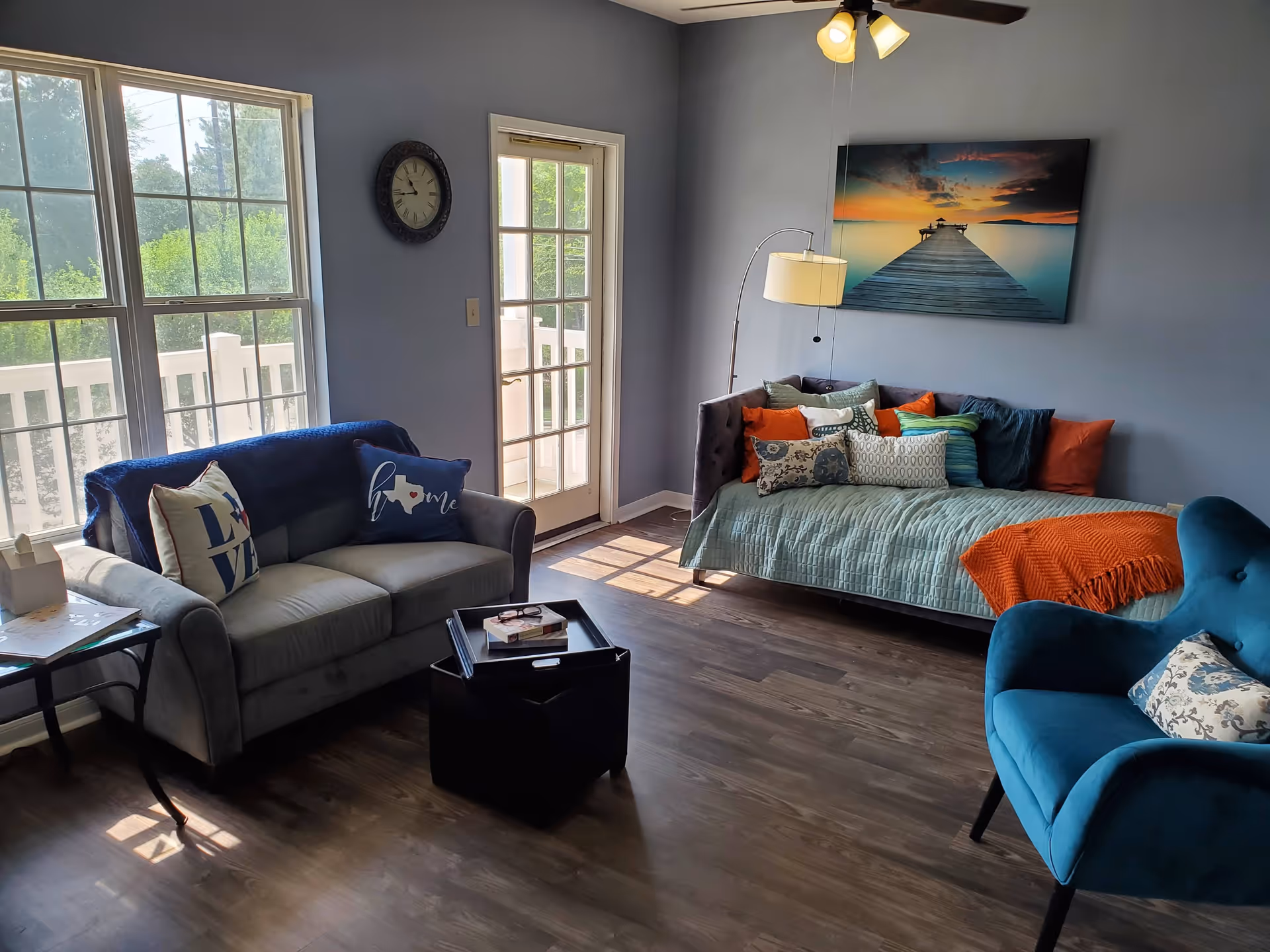 A cozy living room with a gray loveseat adorned with decorative pillows, a blue armchair with a patterned pillow, and a daybed covered with a light blue quilt and multiple colorful pillows. A floor lamp stands next to the daybed, and a large wall clock is mounted above a glass door leading to a balcony. A framed picture of a pier at sunset hangs on the wall. Sunlight streams through large windows, illuminating the wooden floor.