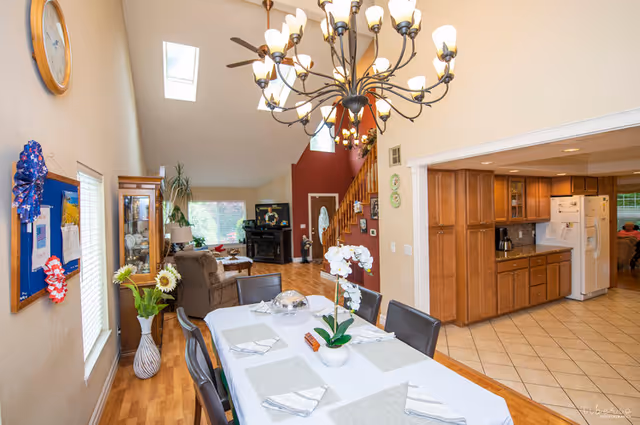 Dining table set beneath a large chandelier opening to a living room with a staircase and adjoining kitchen.