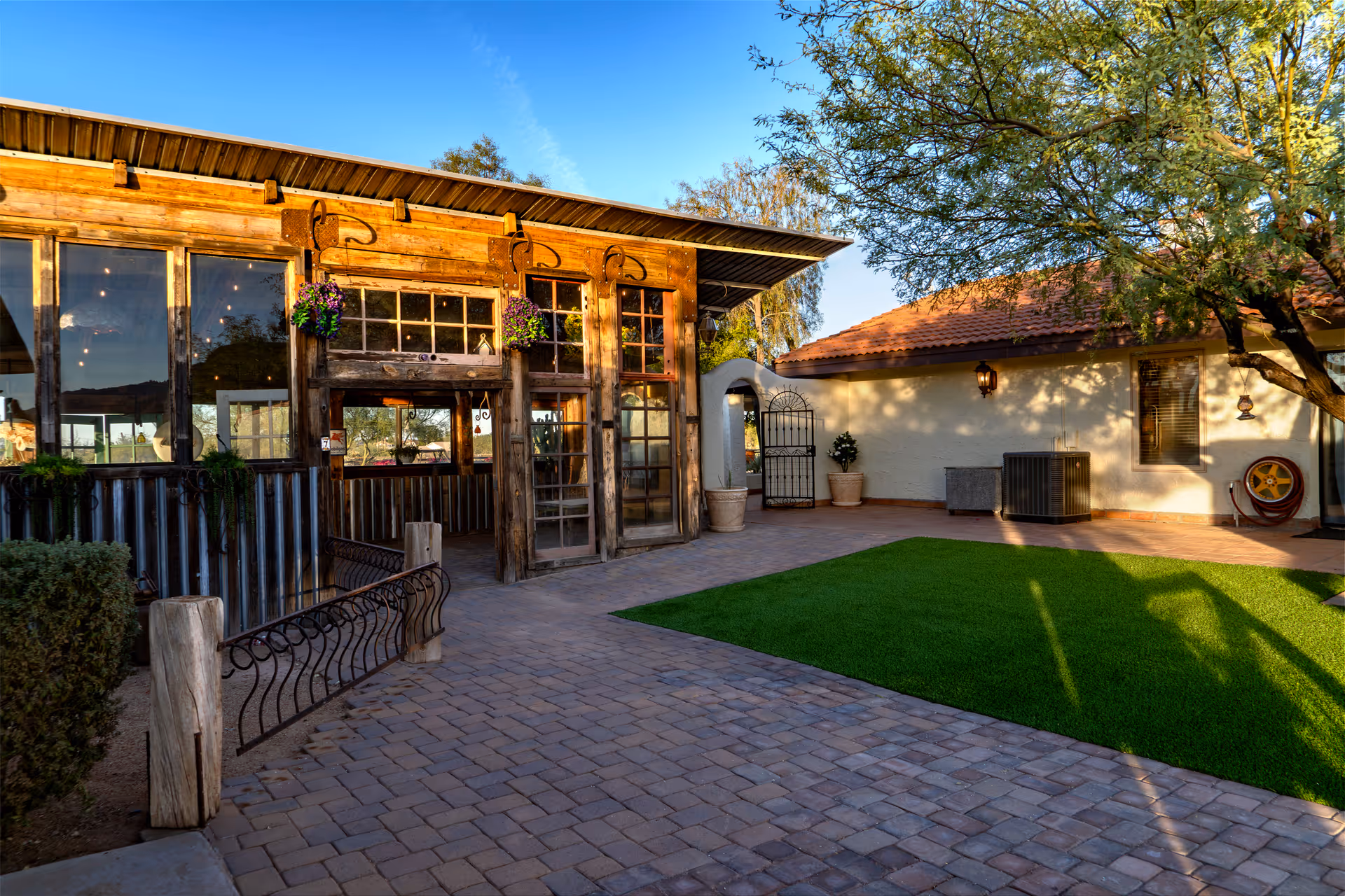 Outdoor courtyard area with a paved stone walkway, green artificial grass, and a rustic wooden building with large windows and hanging flower pots. There is a tree casting shadows on the ground and a white building with a red tiled roof in the background.