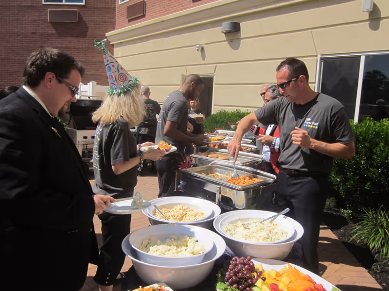 Several people serving themselves food from a buffet table set up outdoors next to a building. The buffet includes bowls of salad, pasta, and fruit, as well as trays of hot food. One person is wearing a party hat, and another person is dressed in a black suit. The setting appears to be a casual outdoor gathering or event.