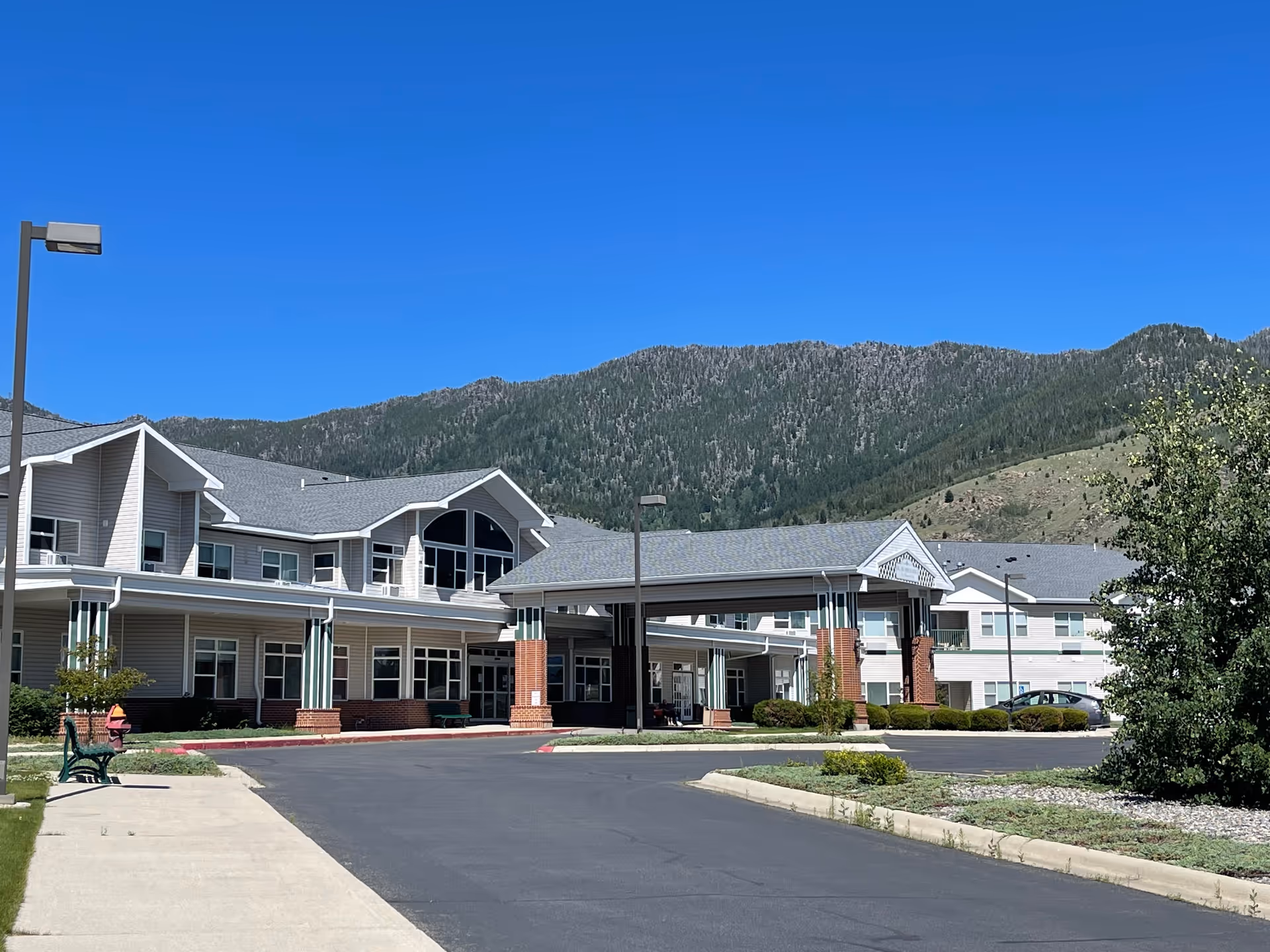 Front entrance of a multi-story senior living facility with a covered porte-cochere, driveway and mountains in the background.