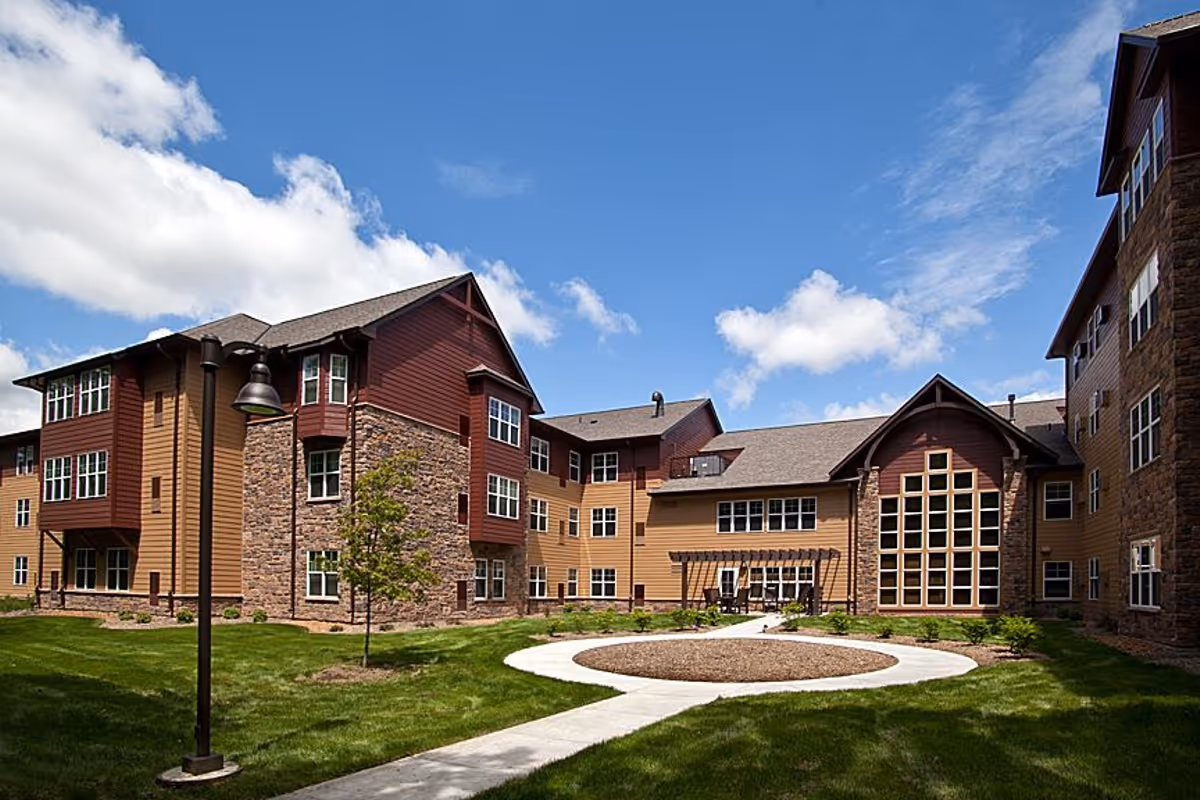 Exterior view of a senior living facility named Valley Ridge, featuring a multi-story building with brown and red siding, stone accents, numerous windows, a well-maintained lawn, a curved concrete walkway, and a clear blue sky with some clouds.