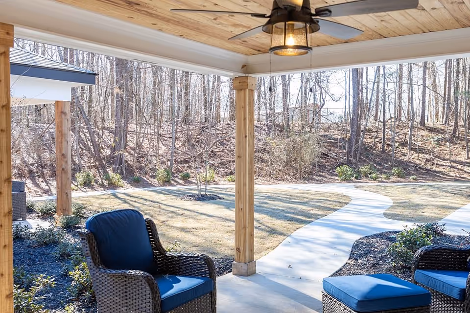 Covered outdoor patio with wicker chairs and blue cushions overlooking a curved walkway and wooded yard.