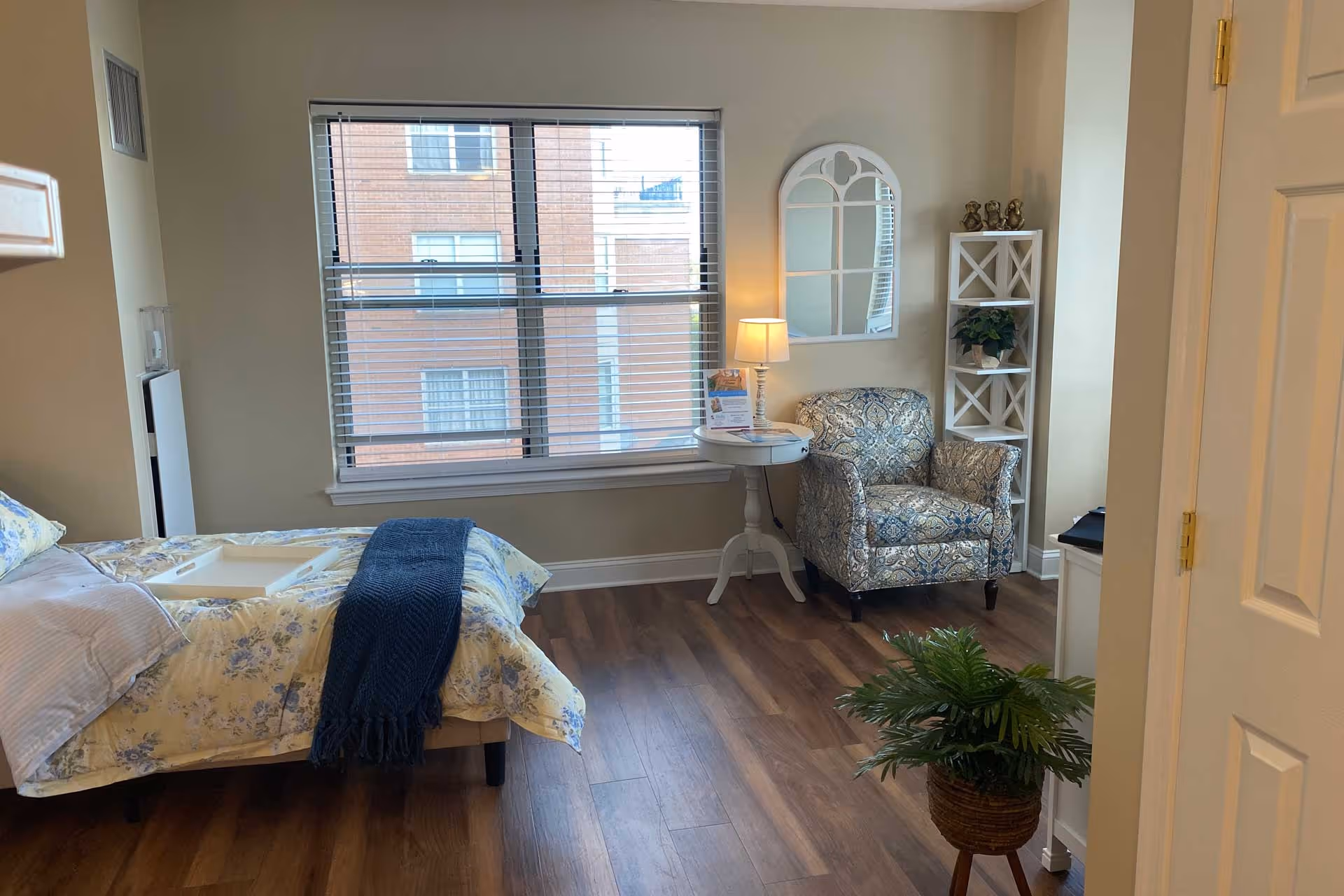 Well-lit bedroom with a bed, patterned armchair, side table with lamp, and a window overlooking a brick building.