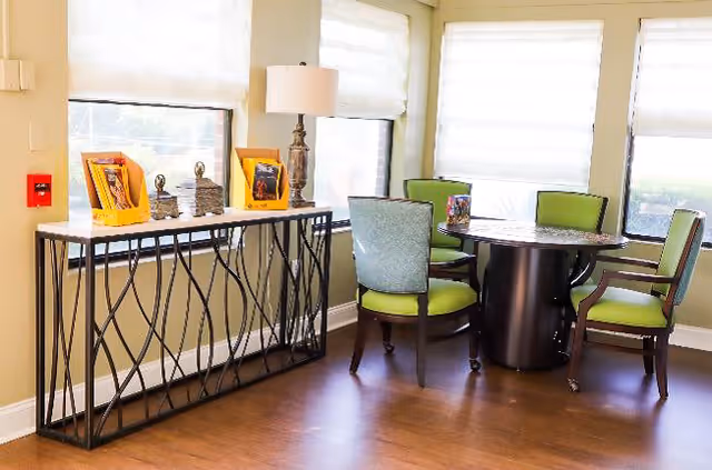 A bright room with large windows covered by white blinds, featuring a round table surrounded by four chairs with green cushions. Against one wall is a decorative console table with a lamp and framed pictures on top. The floor is wooden.