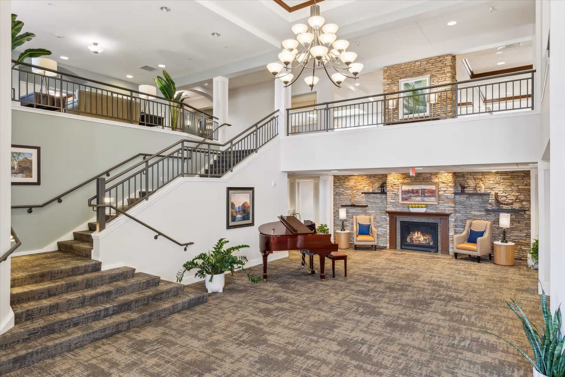 Spacious and well-lit interior common area with a grand piano in the center, carpeted floor, a stone fireplace flanked by two armchairs with blue cushions, and a staircase leading to an upper level with railings and seating areas. The ceiling features a large chandelier and recessed lighting.