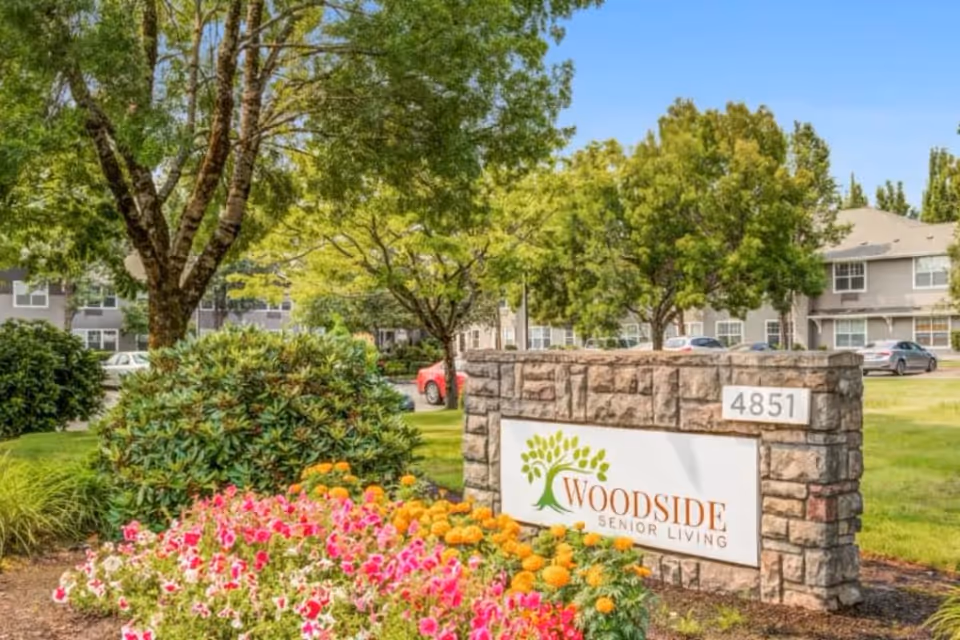 Stone entrance sign reading "Woodside Senior Living" surrounded by flowers and trees with the facility buildings in the background.