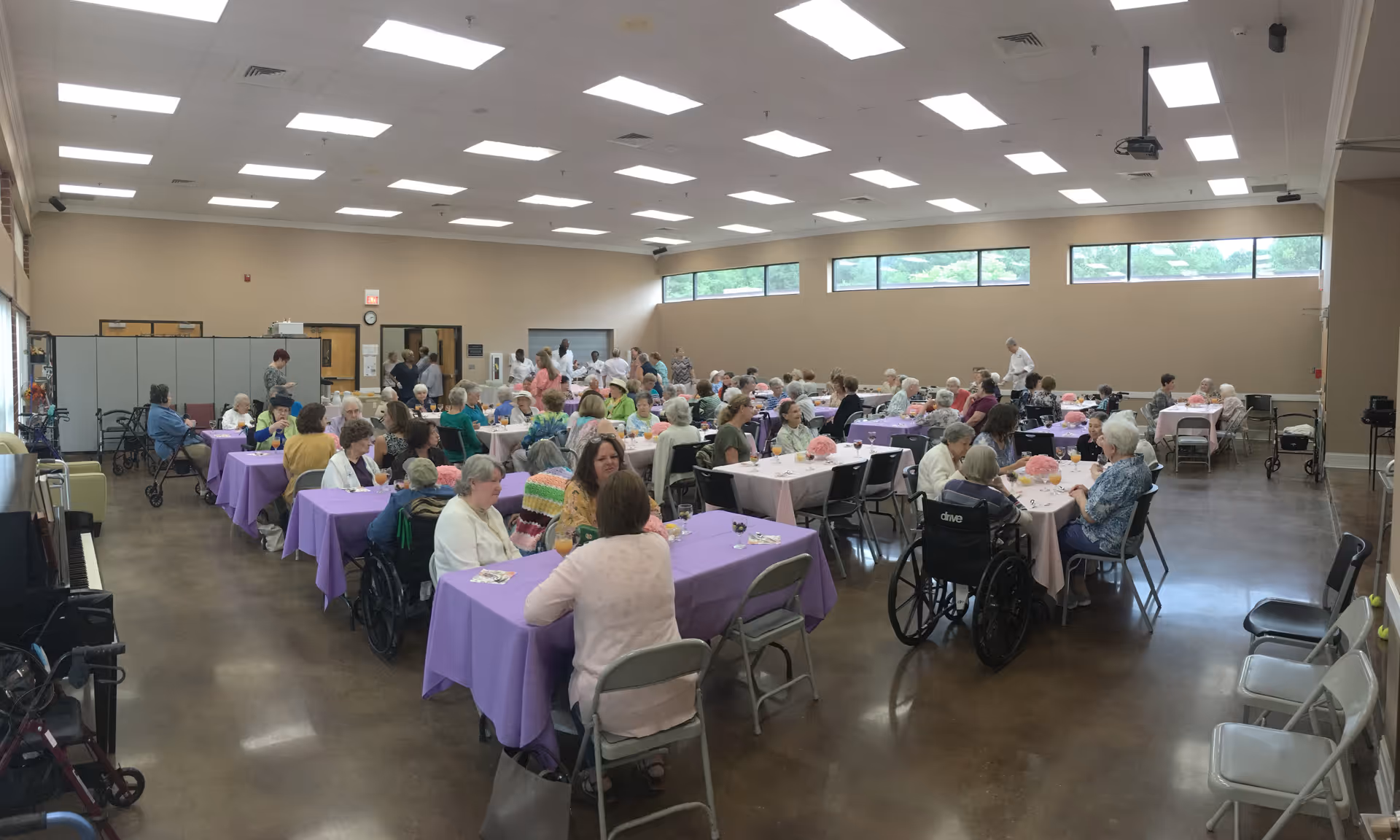 Large community dining/activity hall with many residents seated at tables covered in purple tablecloths.