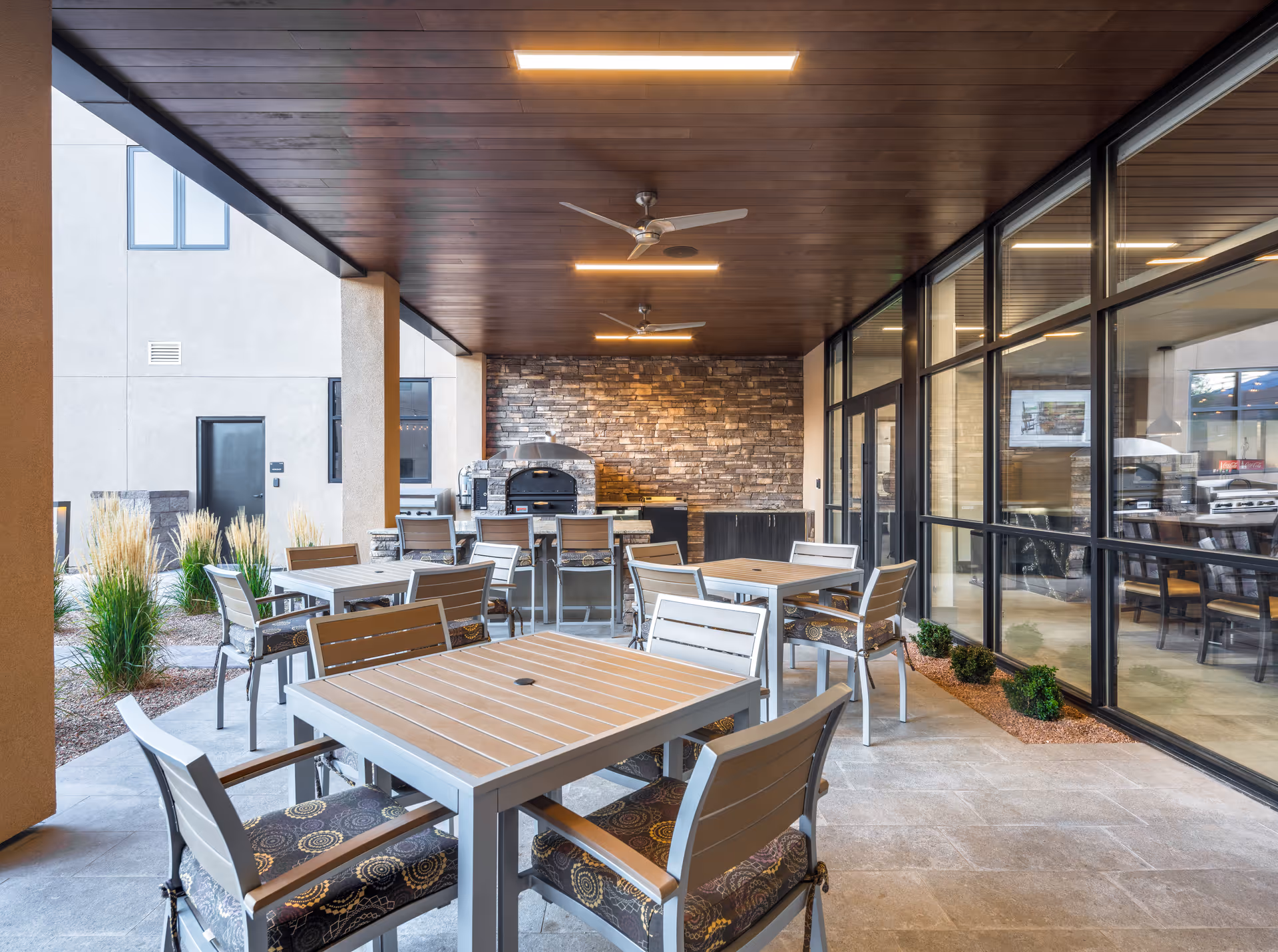 Covered outdoor patio area with multiple tables and chairs, ceiling fans, and a stone wall with a built-in pizza oven and grill. Large windows on one side show an indoor dining area.