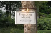 A white sign mounted on a tree trunk with the text 'Friendship Manor Residential Care Assisted Living' surrounded by green grass and trees in the background.