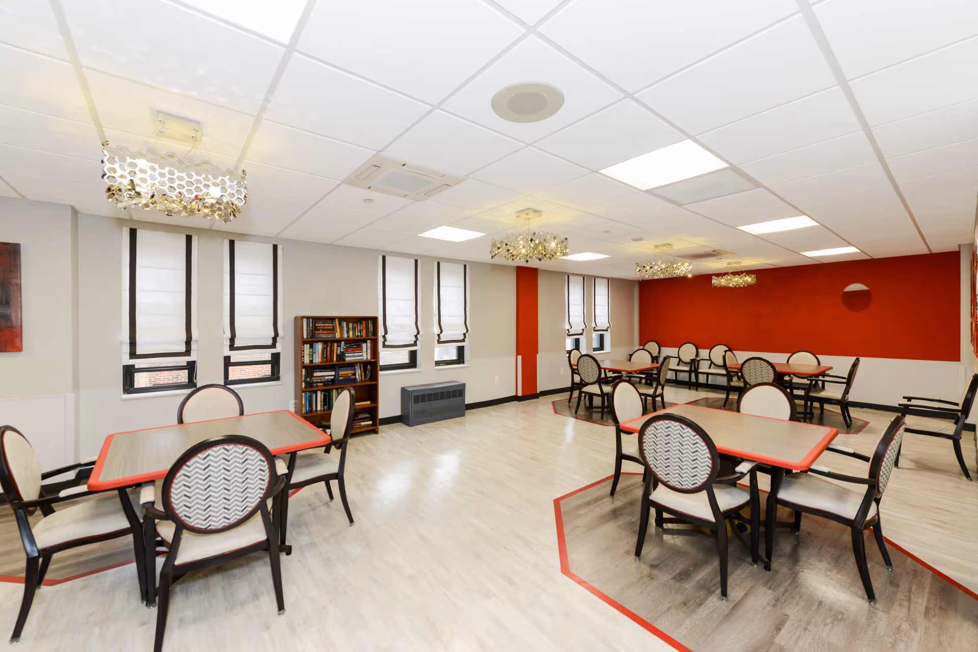 A bright and spacious dining room with multiple tables and chairs arranged neatly. The room features light-colored flooring with red accents around the tables, white walls with a bold red accent wall, several windows with white blinds, and modern ceiling lights. A bookshelf filled with books is placed against one wall.