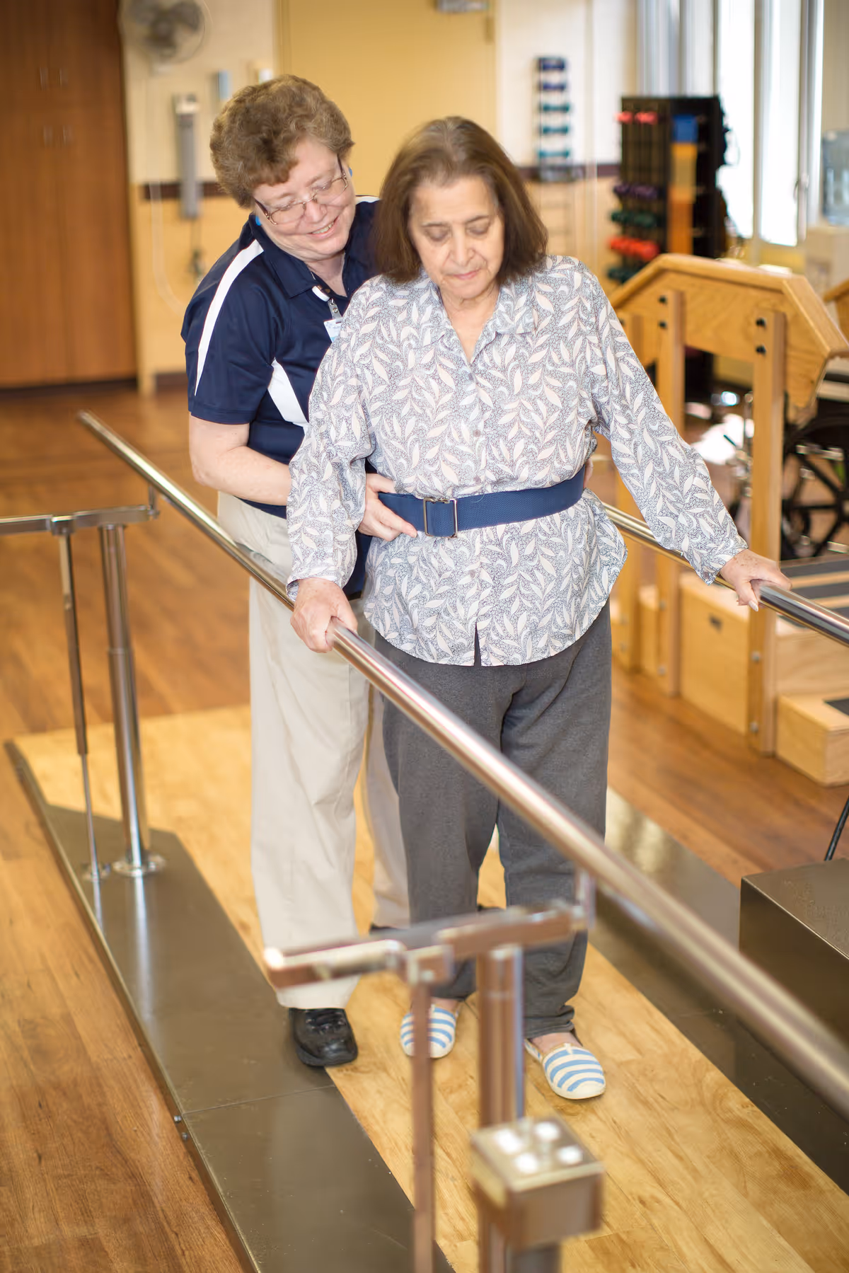 A healthcare worker assists an elderly woman as she walks between parallel bars in a rehabilitation or physical therapy room with wooden flooring and exercise equipment in the background.