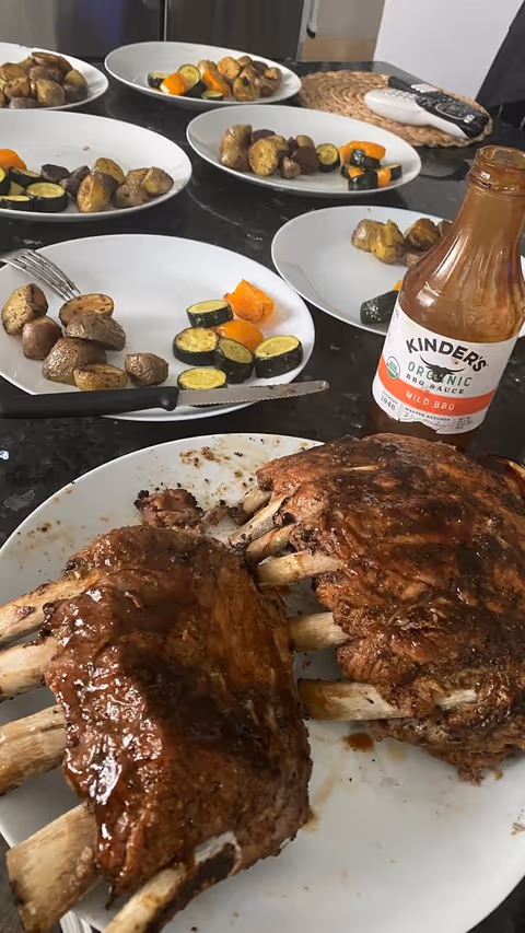 A close-up view of a kitchen counter with plates of roasted vegetables and two racks of barbecued ribs on a white plate. A bottle of Kinder's Organic Mild BBQ Sauce is also visible on the counter along with some utensils and remote controls in the background.