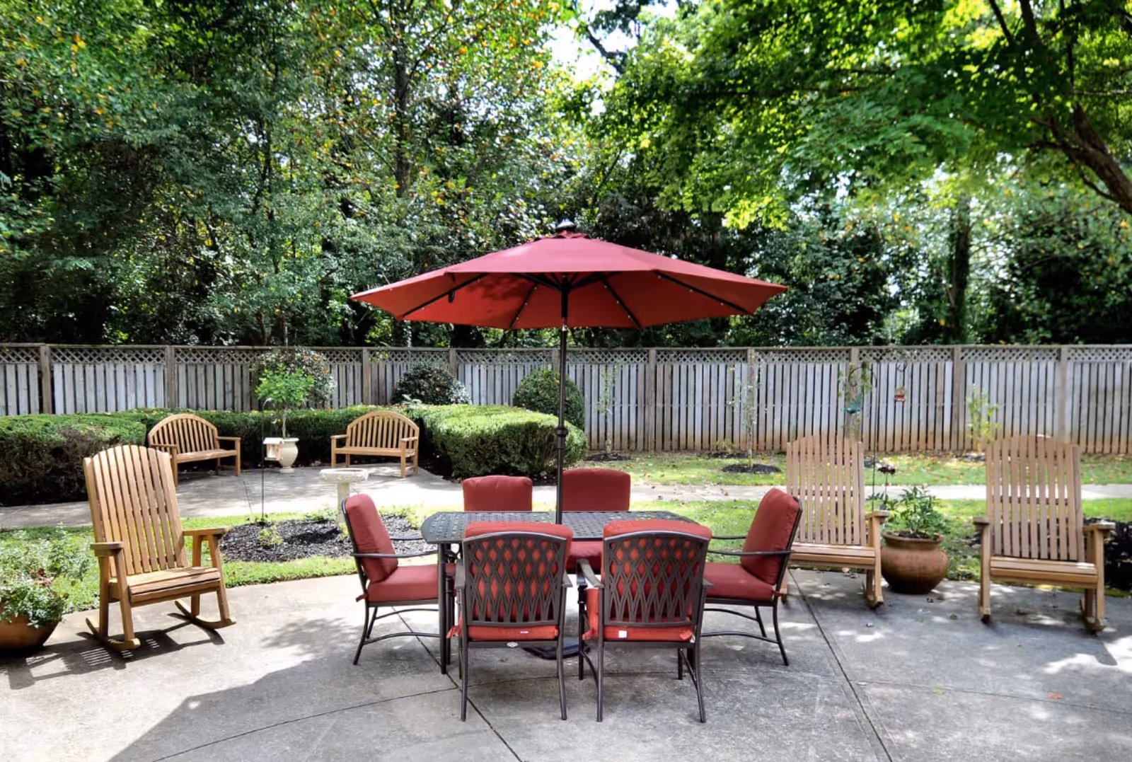 Outdoor patio area with a table and six chairs, all with red cushions, under a large red umbrella. Surrounding the patio are wooden rocking chairs, potted plants, trimmed bushes, and a wooden fence with trees in the background.