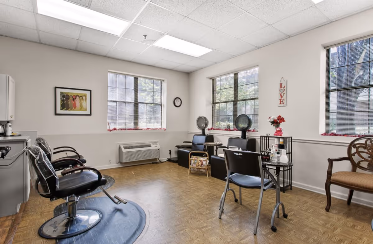 A bright and clean hair salon room with three black salon chairs, two hair dryers, a small table with hair products, and large windows letting in natural light. The walls are light-colored with minimal decorations including a framed picture and a wall hanging that says 'LOVE'.