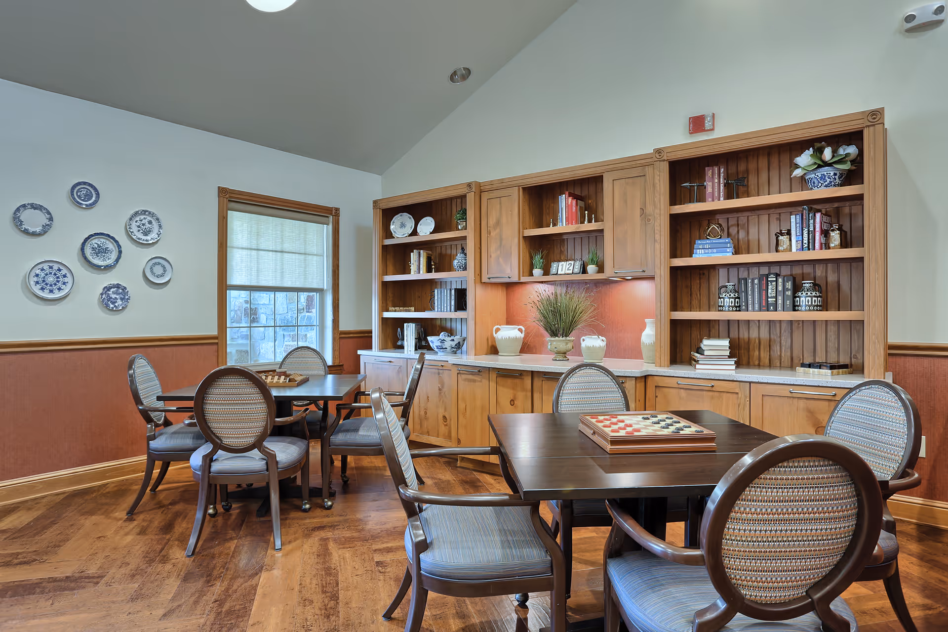 A cozy interior room with wooden flooring and two dark wooden tables, each surrounded by four cushioned chairs with patterned upholstery. One table has a checkers game set up. The back wall features built-in wooden shelves and cabinets decorated with books, vases, and plants. A window with a light shade and decorative blue and white plates on the adjacent wall add to the warm ambiance.