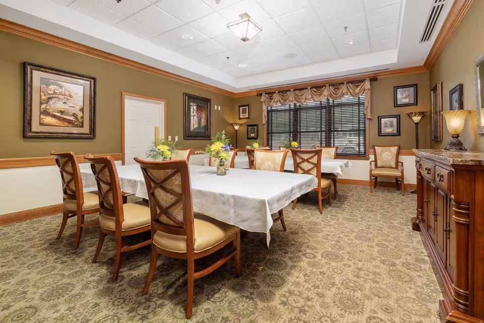 A dining room with two rectangular tables covered with white tablecloths, surrounded by wooden chairs with cushioned seats. The room has patterned carpet flooring, olive green walls with wood trim, framed artwork, and a window with blinds and a valance. There are decorative lamps and a wooden sideboard along one wall.
