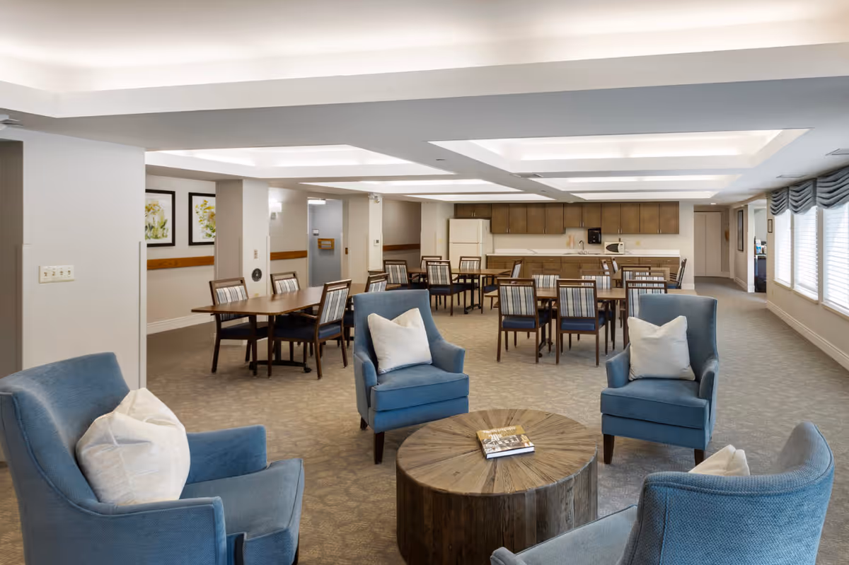 Open senior living common area with blue armchairs around a round wooden coffee table and several dining tables and chairs by a kitchenette.