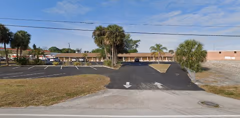 Wide view of the front parking lot and entrance area of Palace Retirement Home, showing a single-story building with a flat roof, palm trees, and a clear blue sky with some clouds.