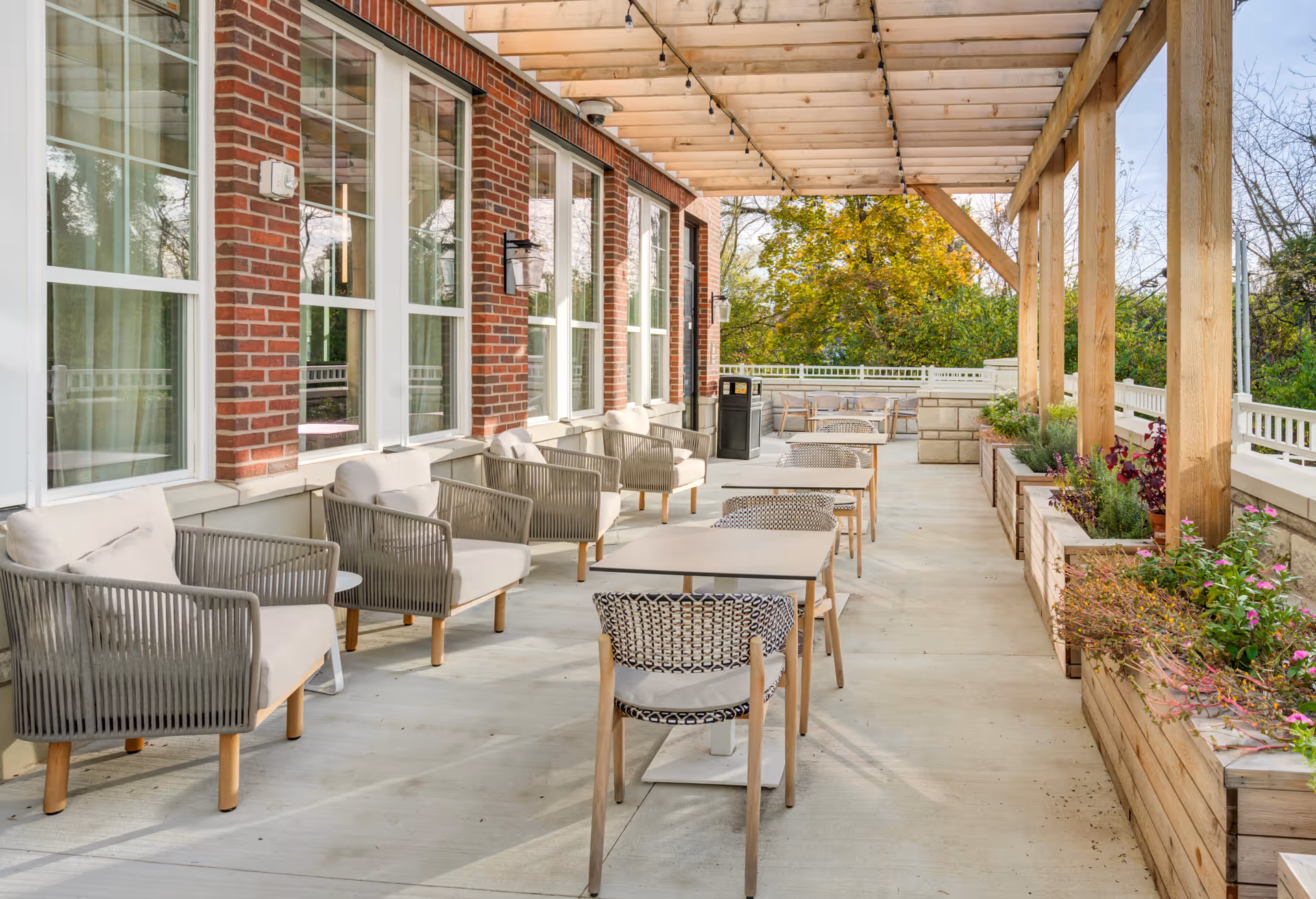 Outdoor patio area at Brookwood Point with cushioned chairs and tables arranged along a brick building under a wooden pergola, surrounded by planters with greenery and flowers.