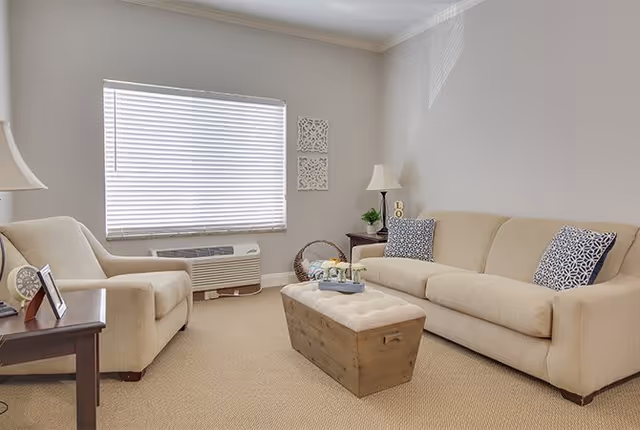 A cozy living room with a beige sofa and matching armchair, a wooden coffee table with a floral centerpiece, a side table with a lamp and picture frame, a window with closed blinds, and neutral-colored walls with decorative wall hangings.
