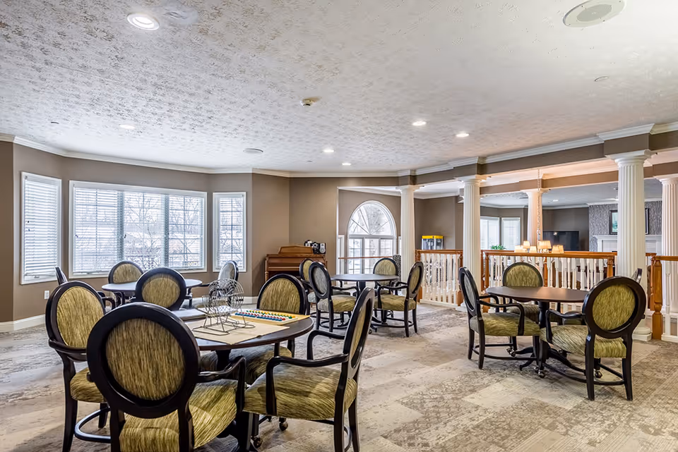 A spacious and well-lit common area with multiple round tables surrounded by green cushioned chairs. Large windows allow natural light to fill the room. The area features beige walls, white columns, and a carpeted floor with a patterned design. A piano is visible in the corner near the windows.
