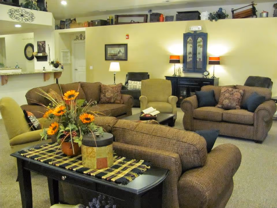 A cozy living room with multiple upholstered sofas and armchairs arranged around a coffee table. There is a decorative table with a floral arrangement featuring sunflowers in the foreground. The walls are painted light yellow, and there are lamps, framed pictures, and decorative items on shelves and tables. The room has a warm and inviting atmosphere.