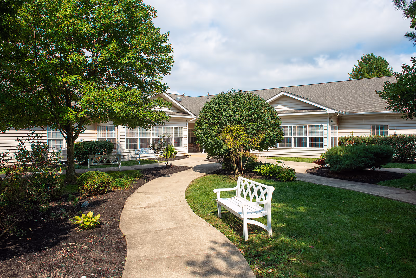 Front courtyard with a curved walkway, benches, and landscaping leading to the entrance of a single-story senior living building.