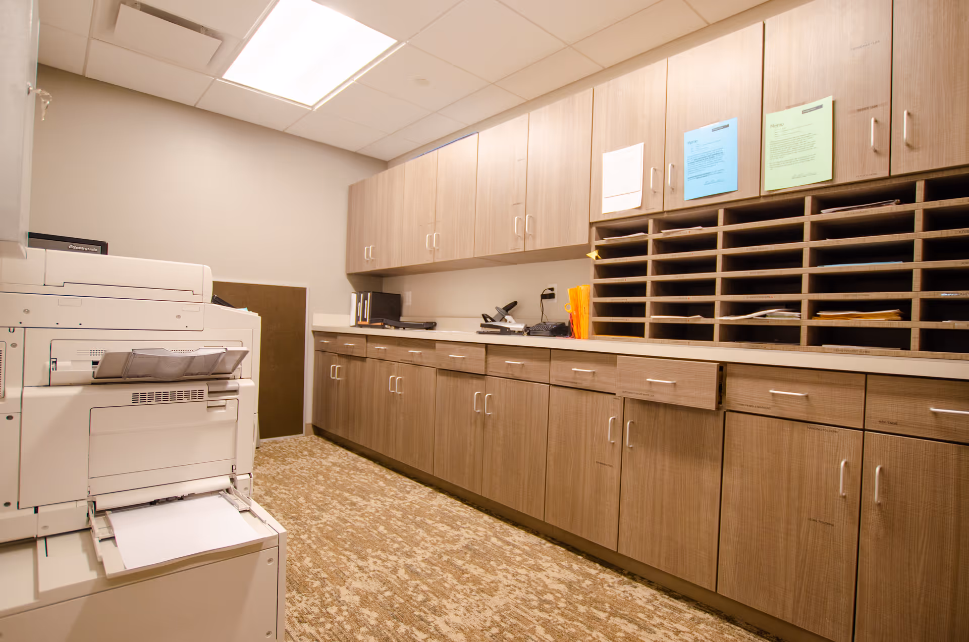 A well-lit interior mail/office room with a large copier, long countertop, numerous wood cabinets and pigeonhole mail slots.
